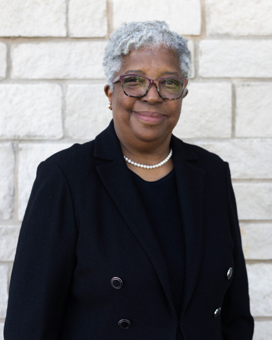 A woman with short hair and glasses standing in front of a brick wall.