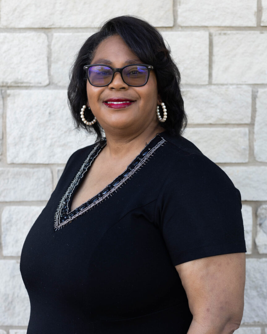 A woman in glasses and black shirt standing next to a wall.