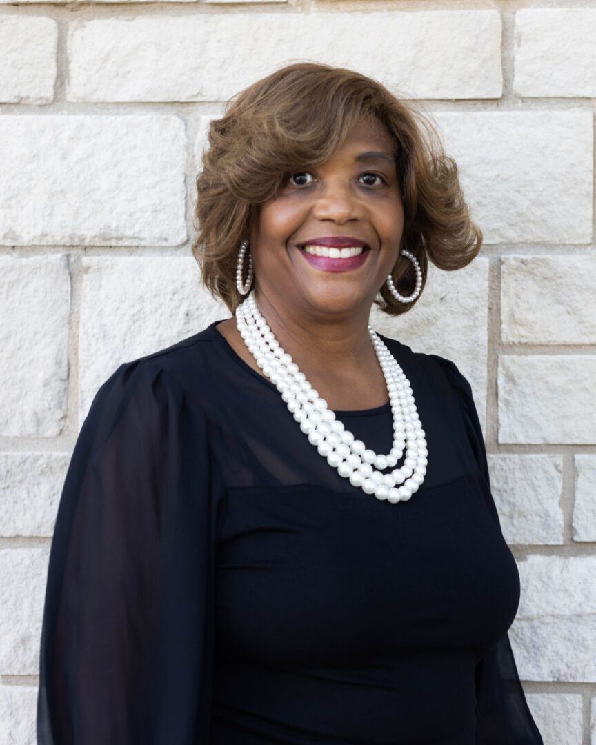 A woman in black dress and pearls standing next to a wall.